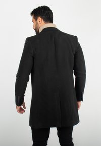Manteau en laine noir avec un design long, des épaules structurées et un col beige. La texture semble lisse, avec des poignets boutonnés.