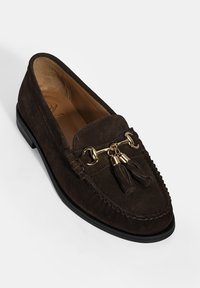 Bruine suede loafer met een ronde neus, decoratieve gouden hardware en franjes. Heeft een gestructureerde bovenkant en een platte rubberen zool.