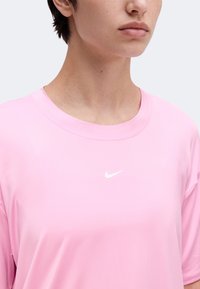 Άτομο που φοράει ένα ανοιχτό ροζ μπλουζάκι Nike με μικρό λευκό λογότυπο Nike στη θωρακική περιοχή, εμφανιζόμενο από τους ώμους έως τα χείλη.