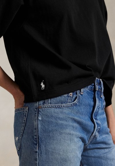 Személy fekete pólót visel, kis fehér póló játékos logóval, világoskék farmernadrágban, keze a hátsó zsebében pihen.