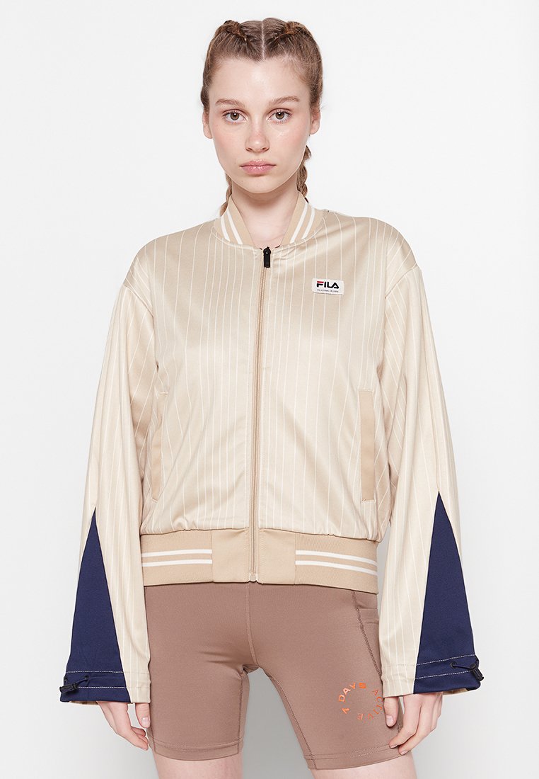 Chaqueta bomber de satén beige con franjas verticales, puños azul marino y cremallera. Logo en el lado izquierdo del pecho; la cintura tiene dos franjas blancas.