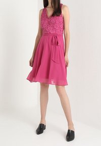 Fuchsiasärks med en blommig spetsliv, V-ringning och en utsvängd chiffongkjol. Utrustad med ett knytbälte och svarta slip-on skor.