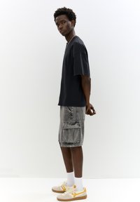 T-shirt noir à manches courtes associé à un short cargo gris clair avec de multiples poches et une coupe décontractée, complété par des baskets beiges.