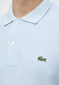 Gros plan d'un polo bleu ciel avec deux boutons blancs et un logo crocodile vert brodé sur la poitrine.