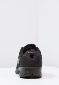 Zwarte sportieve schoen met een bovenwerk van mesh, rubberen zool en een "Quick-Fit" functie aan de achterkant. Het ontwerp is laag en heeft een gestructureerd patroon.