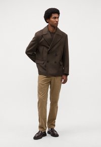 Manteau en laine vert olive à double boutonnage avec des boutons noirs, associé à un pantalon en velours côtelé beige texturé et des chaussures brun foncé.