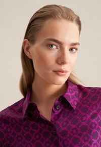 Blusa fucsia con motivo floreale, dotata di colletto con bottoni e bottoni scuri, texture liscia e vestibilità sagomata.