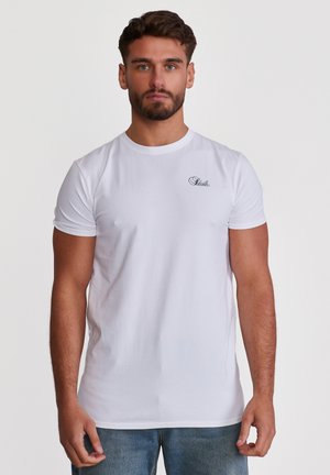 Mladý muž s krátkými tmavými vlasy a vousy, oblečený v jednoduchém bílém tričku s malým černým logem na levé straně hrudi, stojící před bílým pozadím.