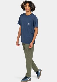 T-shirt in cotone blu navy con maniche corte e taschino sul petto, abbinata a pantaloni verde oliva e sneaker scure con dettagli bianchi.