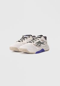 Pár béžovo-bielych športových tenisiek s čiernymi a fialovými akcentmi a textúrovanými béžovými podrážkami na jednoduchom bielom pozadí.