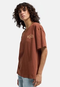 Braunes Baumwoll-T-Shirt mit lockerer Passform, kurzen Ärmeln und einem gestickten Blumenmuster auf der linken Brust. Lässiger und atmungsaktiver Stoff.
