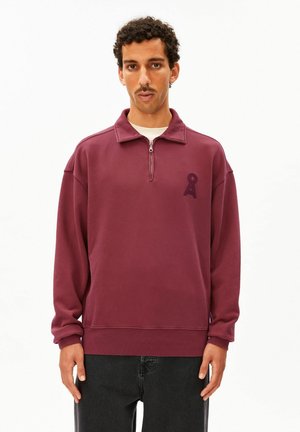 Homme aux cheveux bouclés et à la moustache portant un pull bordeaux à fermeture quart de zip avec un petit logo sombre sur la poitrine gauche et un pantalon noir.