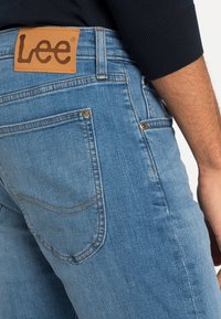 Los jeans de mezclilla azul claro presentan un parche de cuero con el logotipo "Lee". El bolsillo trasero tiene costuras visibles y un pequeño detalle metálico.