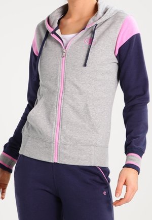 Woman wearing a gray zip-up hoodie with pink accents and navy blue sleeves, paired with navy blue sweatpants with pink details.