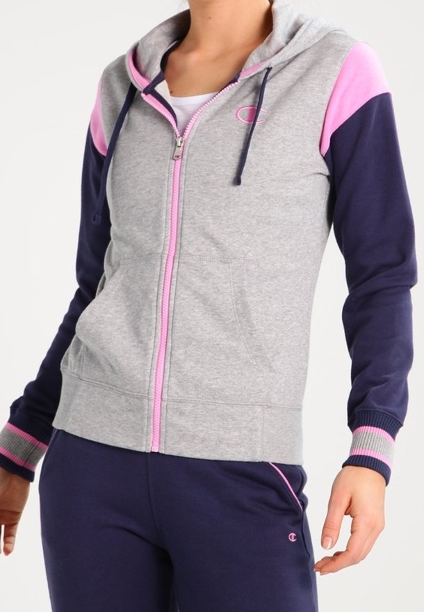 Femme portant un sweat à capuche gris zippé avec des accents roses et des manches bleu marine, associé à un pantalon de survêtement bleu marine avec des détails roses.