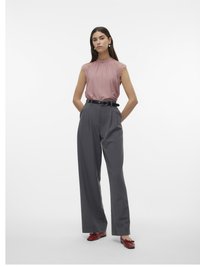 Donna in piedi con le mani in tasca, indossa un top senza maniche in pizzo rosa, pantaloni grigi a vita alta e larghi, una cintura nera e scarpe ballerine rosse.