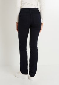 Vaqueros de tiro alto en azul oscuro con pierna ligeramente acampanada, que cuentan con bolsillos traseros y una textura suave. Combinados con zapatillas blancas.