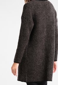 Dlouhý cardigan v černé a hnědé barvě se vzorem herringbone, s kleslými rameny a strukturovaným povrchem, s dlouhými rukávy a uvolněným střihem.