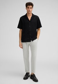 Camicia nera a maniche corte con motivo a chevron testurizzato, abbinata a pantaloni grigi chiari a sigaretta e scarpe nere con lacci.