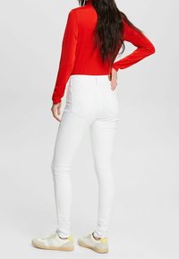 Pantalones vaqueros blancos ajustados con una cintura alta, combinados con una blusa roja ajustada de manga larga y zapatillas ligeras. Fondo liso.