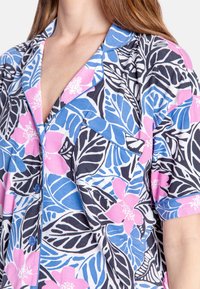 Blommig blus med en blandning av blå och rosa blommor mot ett svartvitt bladmönster; korta ärmar, krage och knappstängning.