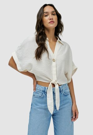 Blusa blanca de manga corta, con botones y un acabado texturizado, con un nudo en la parte delantera. Combinada con unos pantalones vaqueros de cintura alta en un tono azul claro.