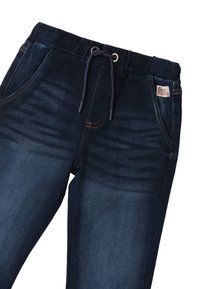 Un pantalon en denim bleu foncé présente une taille à cordon, des poches latérales et une finition texturée subtile. Une étiquette de marque est cousue sur la taille.