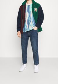 Barevně blokovaná fleecová bunda v tmavě zelené, burgundské a námořnické barvě, pod ní pastelově swirlové tričko, v kombinaci s modrými džíny a bílými teniskami.