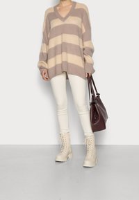 Kvinna iklädd en överdimensionerad beige och taupe randig tröja, vita skinny jeans, krämfärgade snörkängor, och bär en bordeauxfärgad läderhandväska.