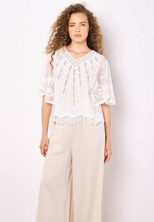 Blusa bianca ricamata con scollo a V, maniche a campana e orlo scallopato; presenta motivi intricati e un tessuto leggero e trasparente.