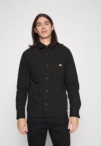 Camisa negra de botones confeccionada con una tela de textura suave, con cuello, dos bolsillos en el pecho y un pequeño parche con el logo.