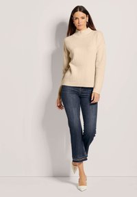 Beiger, gerippter Rollkragenpullover aus Strick, kombiniert mit dunkelblauen Jeans mit geradem Bein und cremeweißen spitzen Pumps. Glatte Textur, maßgeschneiderte Passform.