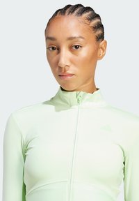 Top zip-up atletico verde chiaro con colletto alto, caratterizzato da un tessuto morbido e accenti a coste. Logo discreto sul lato sinistro superiore.