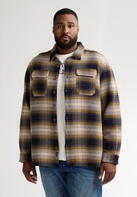 Camicia a quadri in navy, giallo e crema. Tessuto intrecciato con due tasche sul petto e bottoni marroni. Indossata sopra una T-shirt bianca.