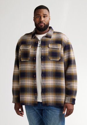 Geruite shirt in marineblauw, geel en crème. Geweven textuur met twee borstzakken en bruine knopen. Gelaagd over een wit T-shirt.
