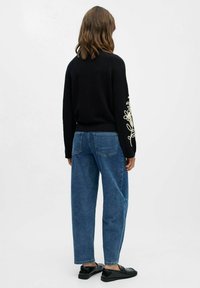 Pull noir avec un motif floral brodé sur la manche gauche, associé à un jean bleu ample et des chaussures noires. Design simple et décontracté.