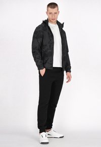Jachetă neagră din nailon cu un model geometric, asortată cu pantaloni sport negri și adidași albi. Modelul stă cu mâinile în buzunare.