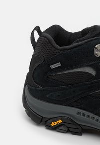 Czarne buty trekkingowe z cholewką z zamszu i siateczki, oznaczone etykietą GORE-TEX, z wyraźną podeszwą Vibram, wzmocnionym piętą oraz perforacjami wentylacyjnymi.