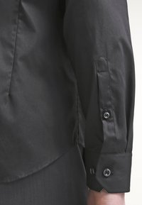 Maglietta nera a maniche lunghe in tessuto liscio con polsini abbottonati, caratterizzata da una leggera lucentezza e da una vestibilità sartoriale. Dettaglio delle spaccature laterali visibile.