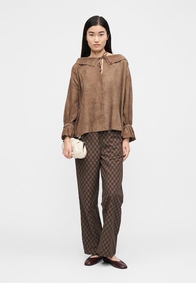 Γυναίκα που φορά χαλαρή καφέ σουέντ μπλούζα με δέσιμο στον λαιμό, καφέ παντελόνι με σχέδιο, κρατώντας μικρή μπεζ τσάντα, όρθια μπροστά σε απλό ανοιχτό φόντο.