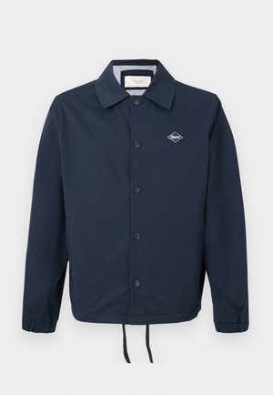 Marineblå jakke laget av glatt stoff, med spiss krage, knapper foran, elastiske mansjetter og snorjustering i kanten med en logo-patch.