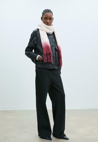 Giacca in pelle nera, pantaloni neri larghi e una sciarpa rosa chiaro con effetto ombre bordeaux e dettagli a frange, su uno sfondo neutro.