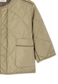 Veste beige matelassée avec couture en forme de losange, dotée d'une grande poche avant et de manches trois-quarts, fabriquée en tissu rembourré.