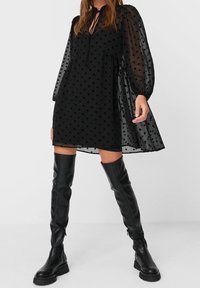 Zwarte jurk met stippen, lange doorzichtige mouwen, getailleerd in de taille en met een strik rond de hals. Gecombineerd met kniehoge zwarte leren laarzen.