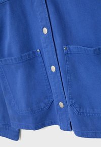 Devant d'une chemise en tissu bleu avec deux grandes poches carrées et des boutons-pression argentés au centre.