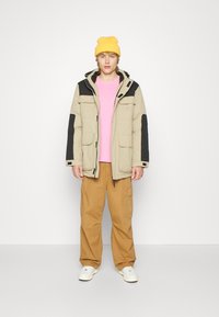 Beige parka-jakke med svarte detaljer, rosa skjorte, beige cargo-bukser, gul lue og hvite joggesko; flere lommer på jakken.