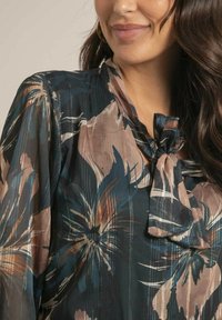 Blouse à imprimé floral en vert canard, avec un col à nouer. Tissu doux et transparent avec une texture lisse et des fils métalliques subtils tout au long.