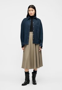 Vrouw staand, gekleed in een marineblauwe denim jas over een zwarte coltrui, beige midirok, zwarte sokken en zwarte leren laarzen tot halverwege de kuit, tegen een witte achtergrond.