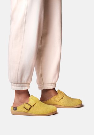 Fødder i gule slip-on tøfler med spænder, båret med lysebeige joggingbukser mod en ensfarvet baggrund.