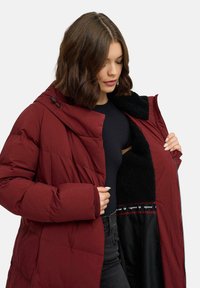 Giacca imbottita bordeaux con cappuccio, dotata di chiusura con zip e fodera in pile nero testurizzato. Include polsini a coste e un dettaglio del logo interno.